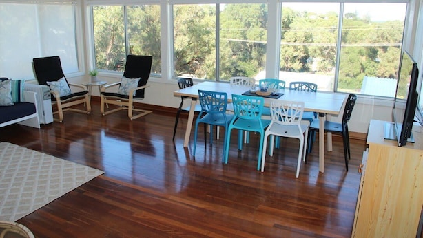 Una habitación amplia con piso de madera, una mesa de comedor rodeada de sillas y grandes ventanales con vistas a árboles.