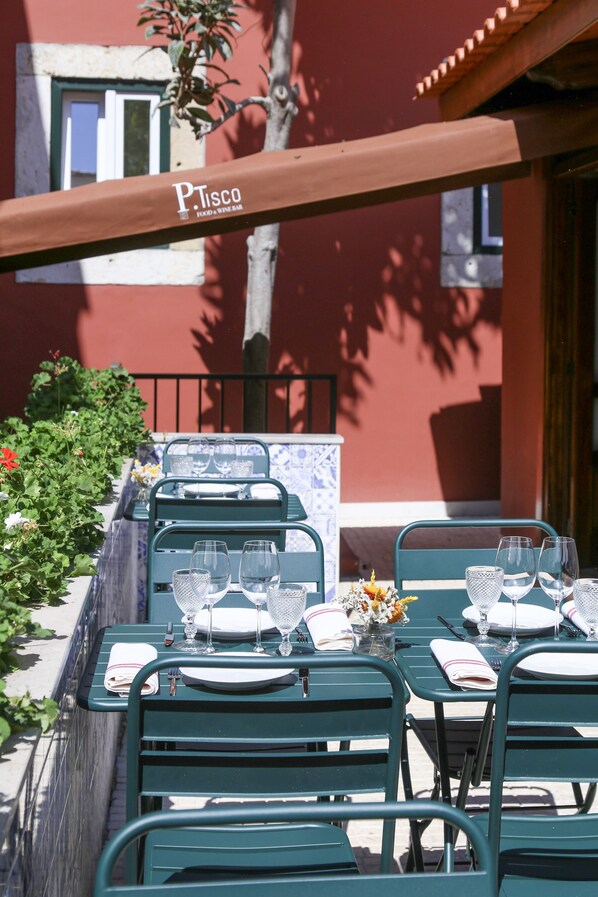 Un área de comedor al aire libre con mesas y sillas de metal verde, cada conjunto con un plato blanco, varios vasos de vino y cubiertos.