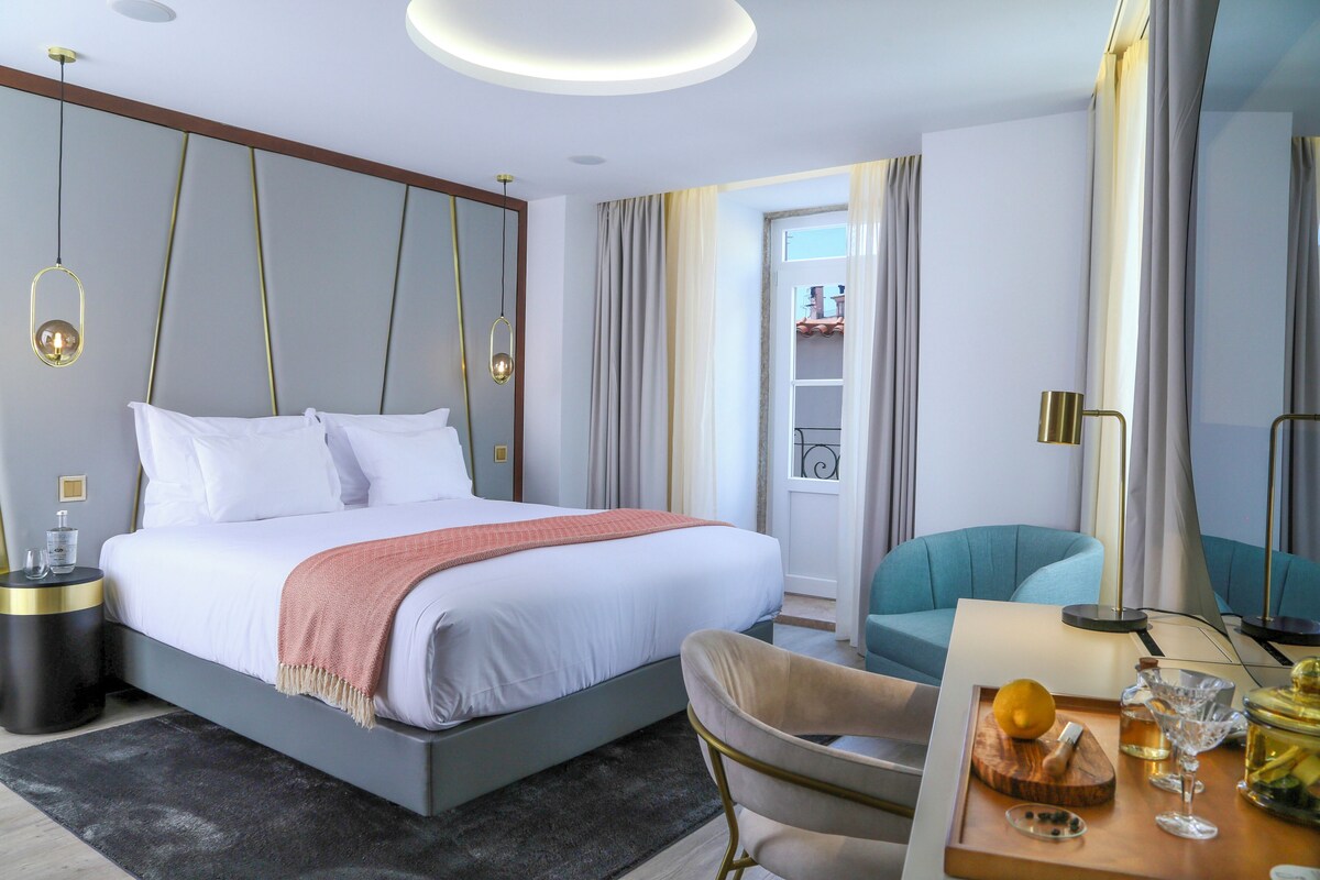 A modern hotel room with a large bed, a desk with a fruit bowl, and a chair.