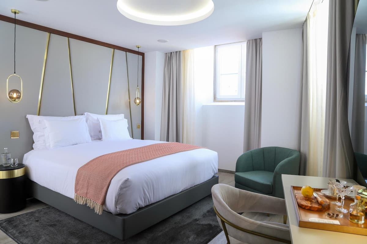 Una habitación de hotel moderna con una cama grande, un sillón verde, una mesita con una bandeja de comida y bebidas, y un ventanal con cortinas.