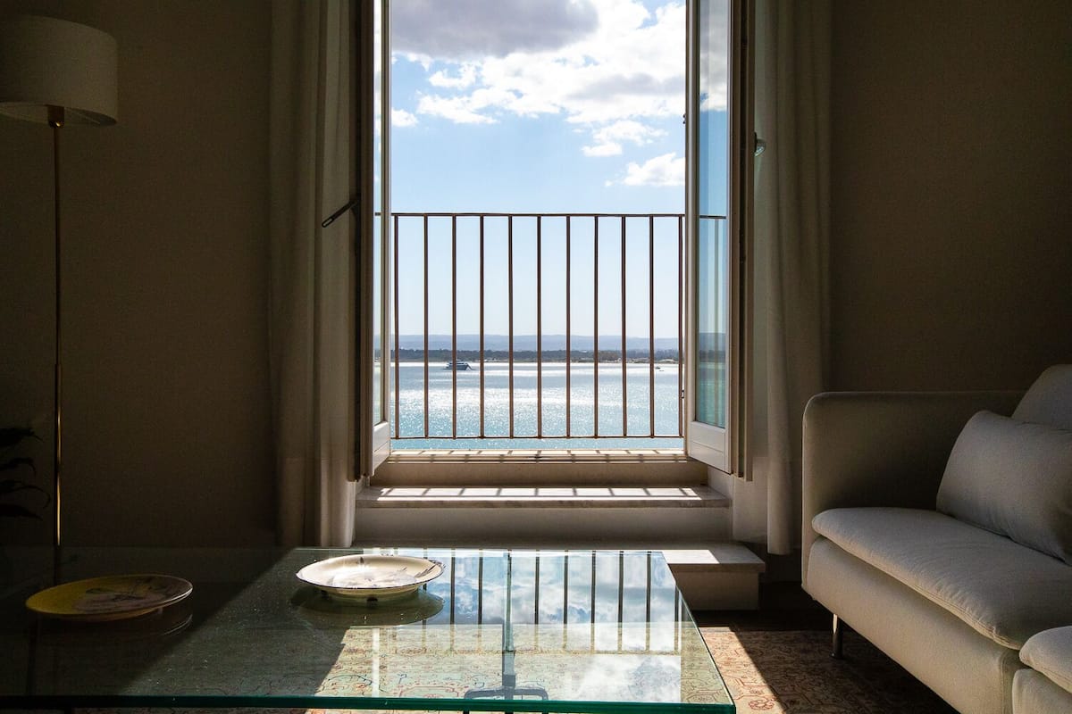 A room with a view of the sea through a window with a metal railing, a sofa, a small table with a plate, and a lamp.