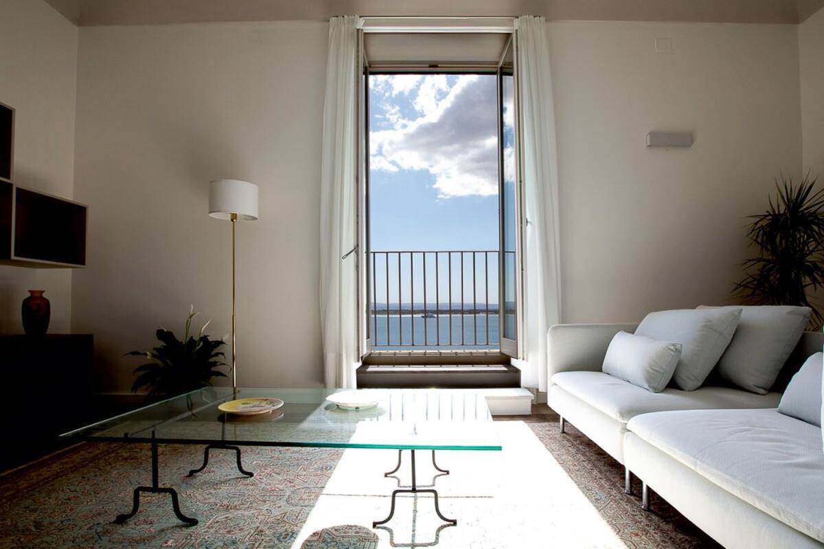 A living room with a glass coffee table, a white sofa, a floor lamp, and a view of the sea through a balcony.