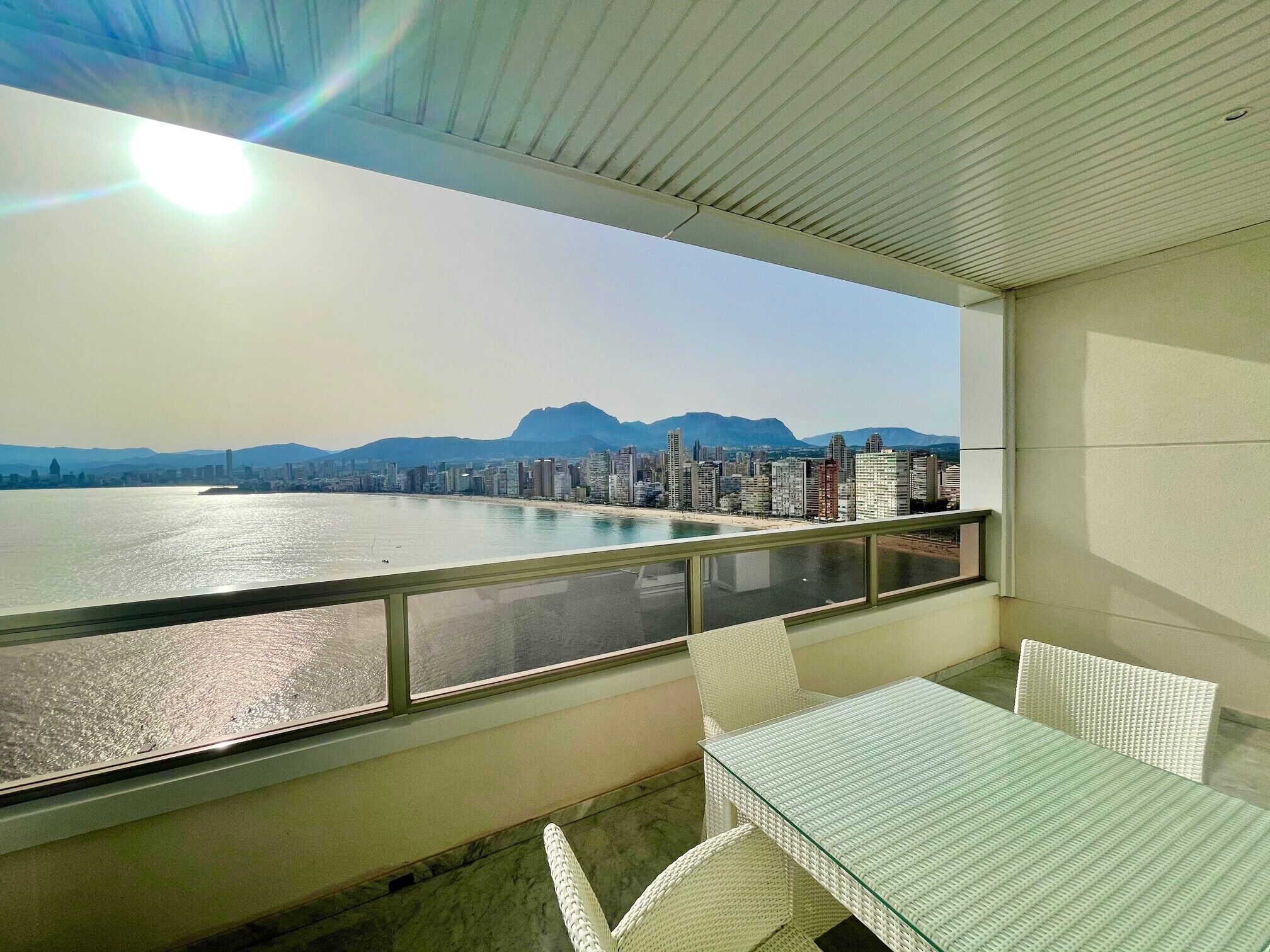 A balcony with a glass table and chairs, overlooking a cityscape and a body of water.