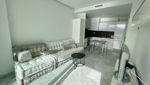A modern living room with a sofa, a small table, and a TV. There is a kitchen area with cabinets and appliances in the background. - MEDITERRANEAN VIEW BENIDORM R078 (Benidorm)