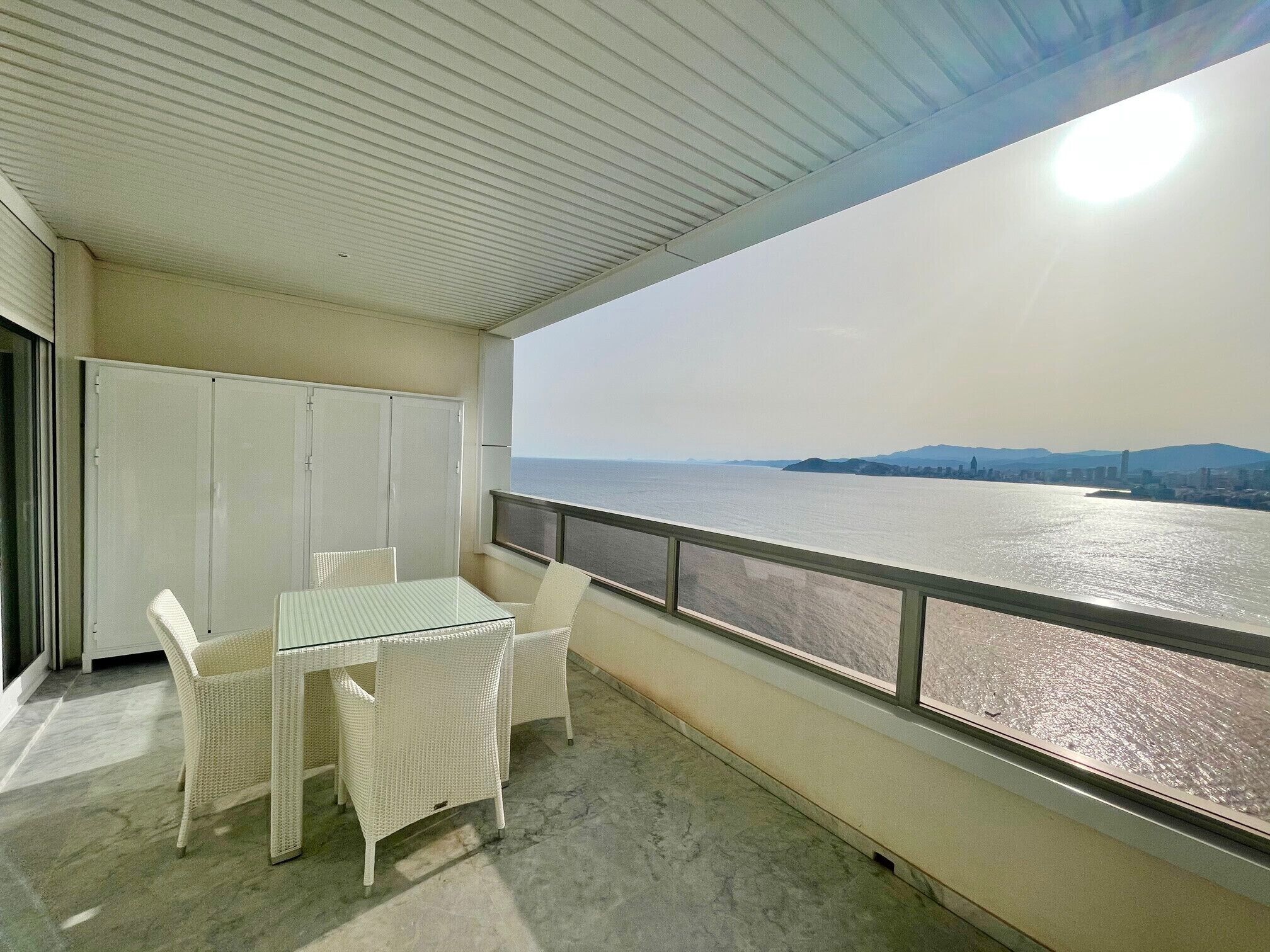 A balcony with a table and chairs, a view of a body of water, and a distant cityscape.