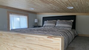 A bedroom with a wooden headboard, a large bed with striped bedding, a nightstand with a lamp, and a window with blinds. - Cozy Tiny Home in Heart of Big Sandy (Big Sandy)