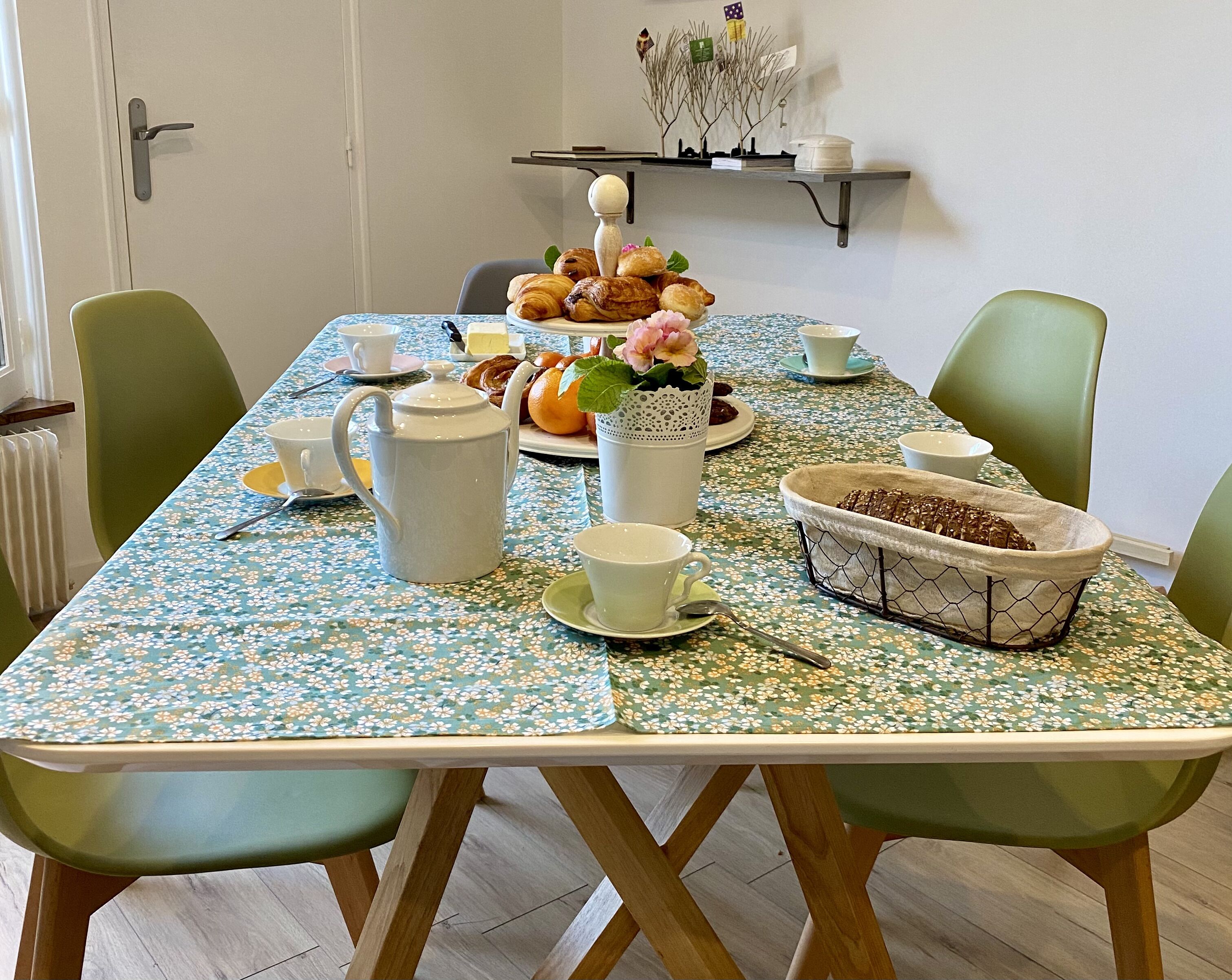 Een eettafel met een tafelkleed met bloemen, een theepot, kopjes, een mand met brood en een vaas met bloemen.