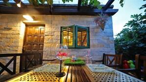 Une table en bois placée à l’extérieur, avec un verre et une bougie, adossée à un mur de pierre. On aperçoit une porte en bois et une fenêtre avec des volets verts.