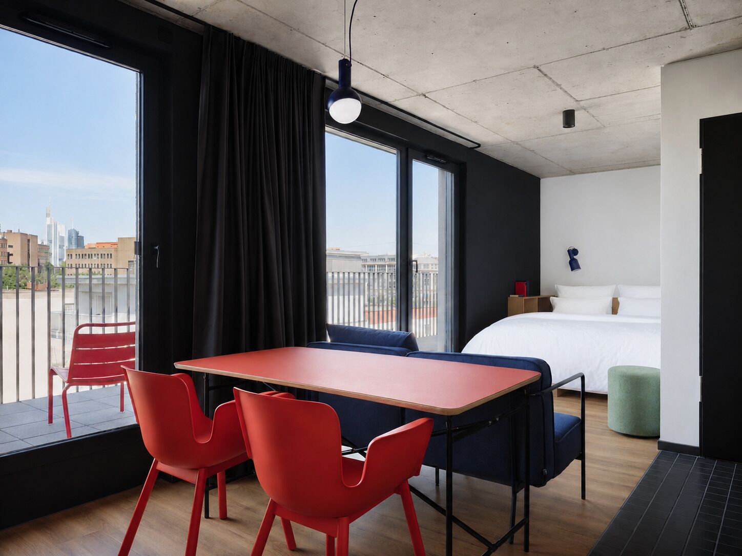 Una habitación de hotel moderna con una cama grande, una mesa de comedor con sillas, un balcón con una silla roja y vistas a la ciudad.