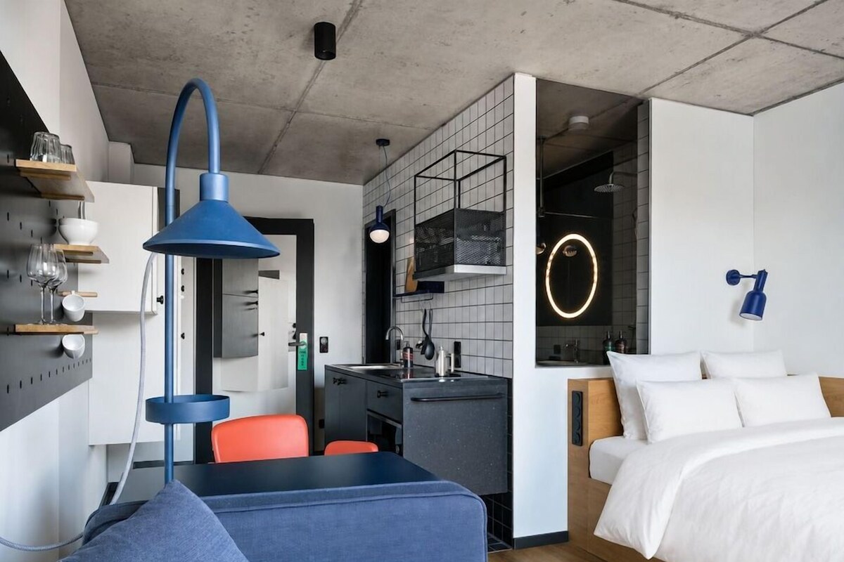 A modern hotel room with a bed, a desk, a chair, and a kitchenette.