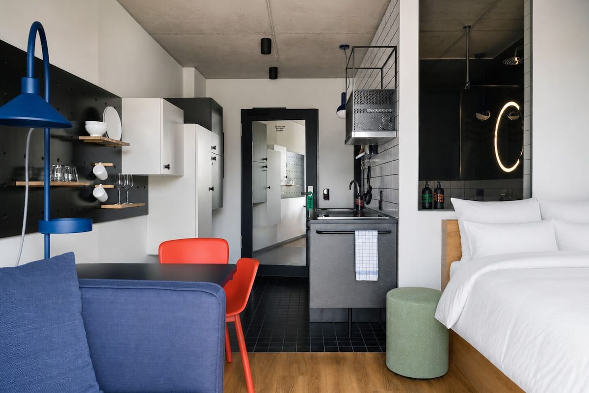 A modern hotel room with a sofa, a bed, a kitchenette, and a bathroom.