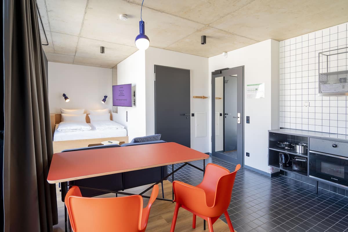 A modern hotel room with a bed, a dining table with chairs, a kitchenette, and a bathroom visible through an open door.