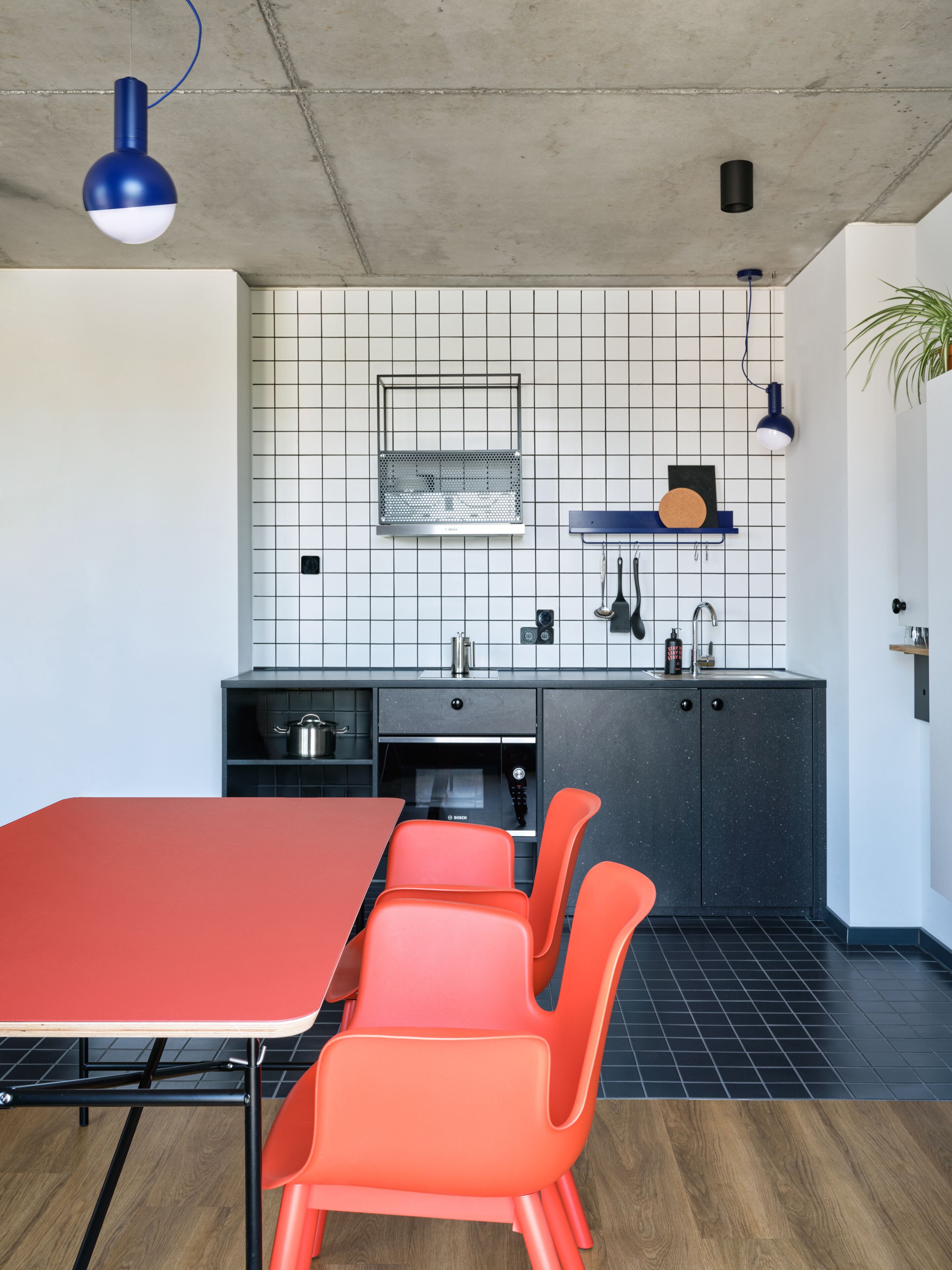 A modern kitchen with a red table and orange chairs, a black countertop, a sink, and a backsplash with a grid pattern.