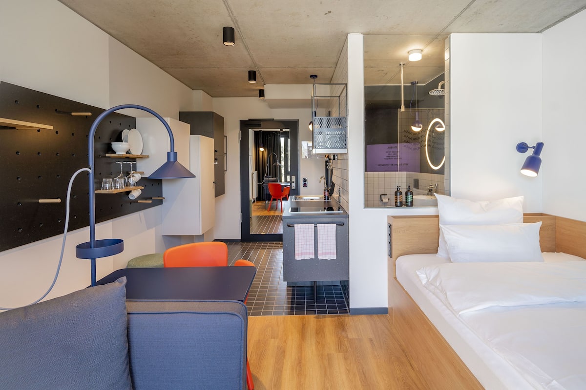 A modern hotel room with a bed, a desk, a chair, a small kitchenette, and a bathroom visible through an open door.