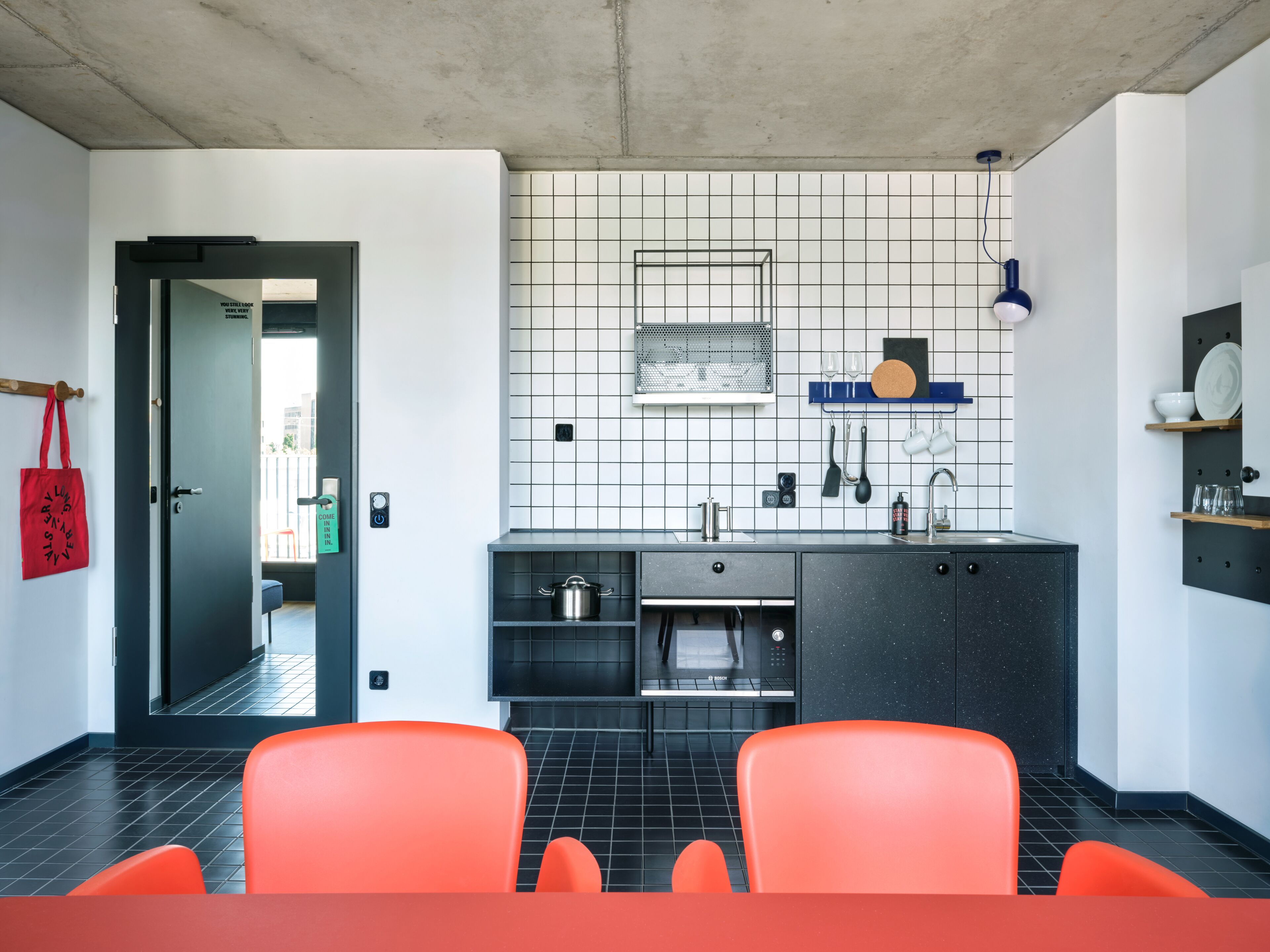 Eine moderne Küche mit einem Bosch-Ofen, einer Spüle und einer gefliesten Spritzschutzwand. Im Vordergrund stehen orangefarbene Stühle und an der Wand hängt eine rote Tasche.