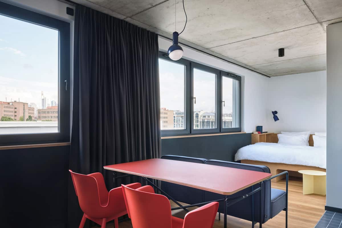 A modern hotel room with a large window, a bed, a desk with chairs, and a city view.