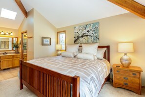 A bedroom with a wooden bed, a nightstand with a lamp, a painting on the wall, and a bathroom with a mirror and sink.