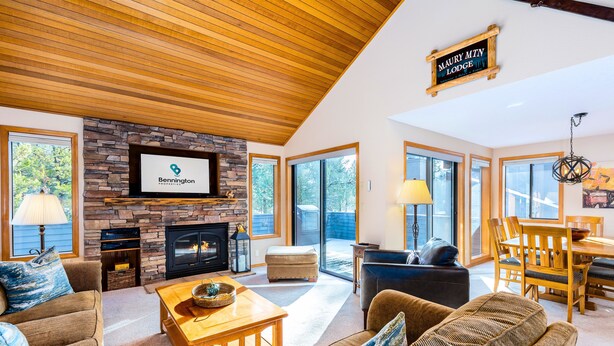 A spacious living room with a stone fireplace, comfortable seating, and large windows offering views of the outdoors. - Maur32-North Sunriver Retreat | Hot Tub, AC, Bikes, Ping Pong (Sunriver)