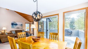 A dining area with a wooden table and chairs, a hanging light fixture, and a view of a deck through large sliding glass doors. - Maur32-North Sunriver Retreat | Hot Tub, AC, Bikes, Ping Pong (Sunriver)