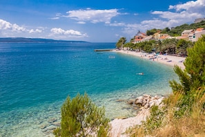 A coastal beach scene with clear waters, a sandy shore, and buildings in the background.