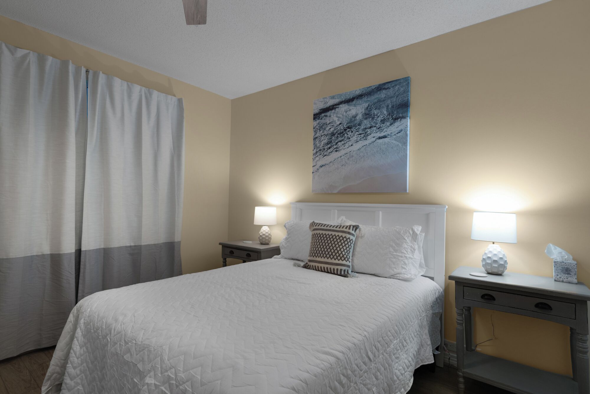 Een hotelkamer met een bed, twee nachtkastjes met lampen, een groot raam met gordijnen en een schilderij aan de muur.