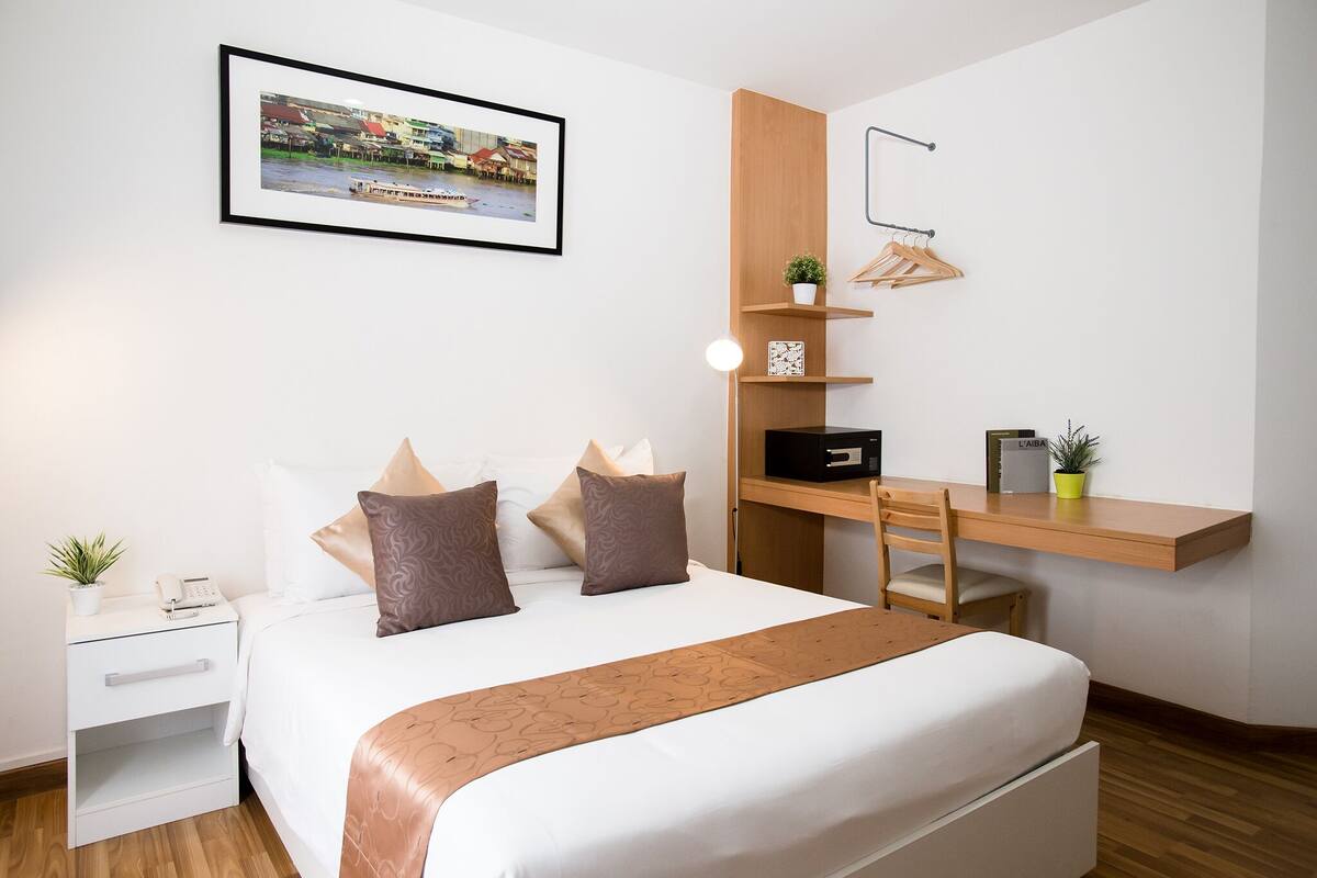 A neatly arranged hotel room with a bed, bedside table, desk, and a wall-mounted shelf with a plant and a framed picture.