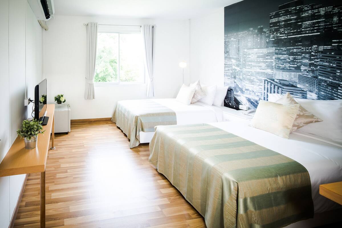 A hotel room with two beds, a desk with a computer, a window with curtains, and a cityscape mural on the wall.