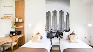 A hotel room with two beds, a desk with a chair, a shelf with a vase and a plant, and a cityscape wall art. - Anda Bangkok Hotel (Bangkok)