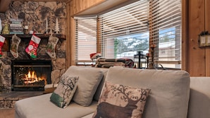 A cozy living room with a fireplace, a sofa with decorative pillows, and a window with blinds. - Walk to Bear Resort with Slope Views. Close to Zoo & Golf, Spa, BBQ & Arcade! (Big Bear Lake)