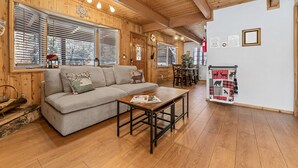 A cozy room with a wooden ceiling, a sofa, a coffee table, and a dining area with a table and chairs. - Walk to Bear Resort with Slope Views. Close to Zoo & Golf, Spa, BBQ & Arcade! (Big Bear Lake)
