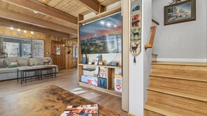 A cozy living room with a large wooden table, a sofa, and a TV mounted on the wall. - Walk to Bear Resort with Slope Views. Close to Zoo & Golf, Spa, BBQ & Arcade! (Big Bear Lake)