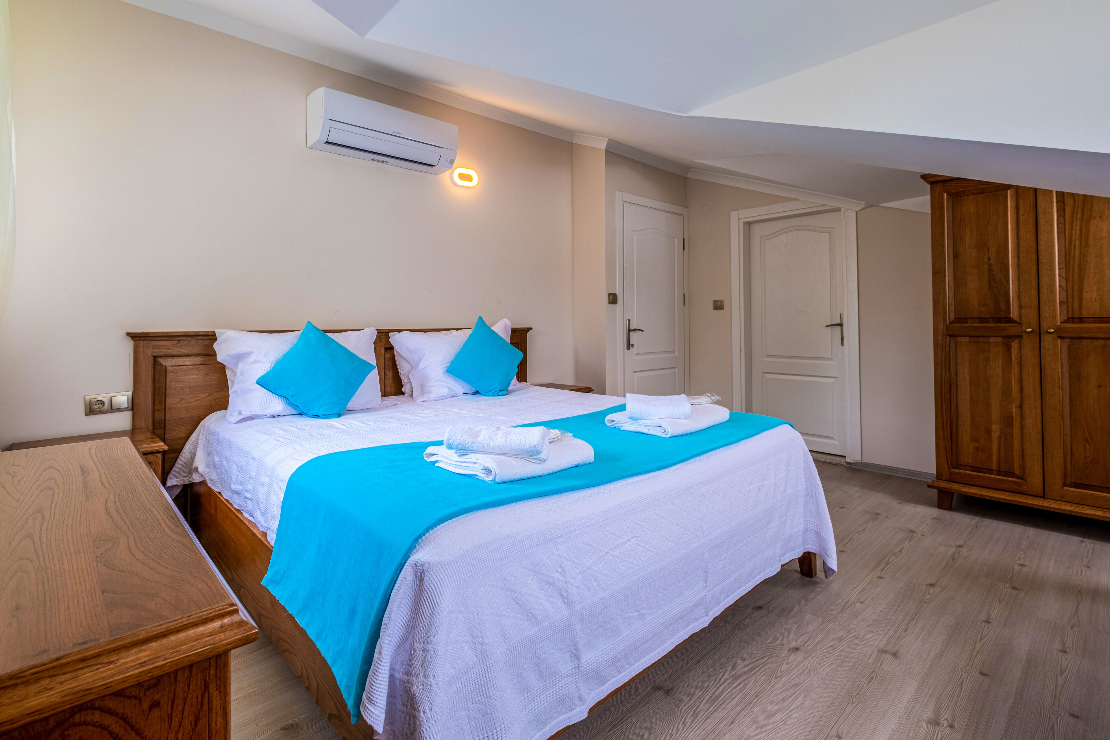 Une chambre d’hôtel avec un lit en bois, du linge de lit blanc, une touche de couleur bleue, une table de chevet en bois, un climatiseur et une armoire.