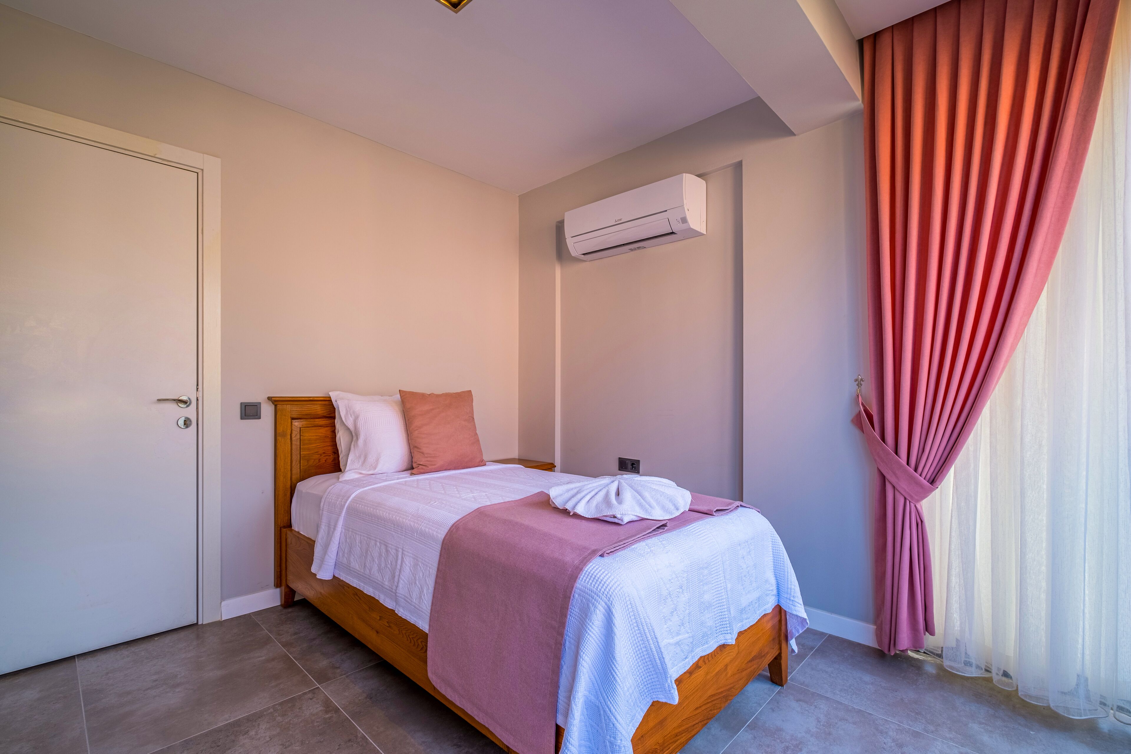 A neatly made bed with a wooden frame, a white and pink bedspread, and a white pillow. There is a white air conditioning unit on the wall, and a window with sheer white curtains and a pink curtain.