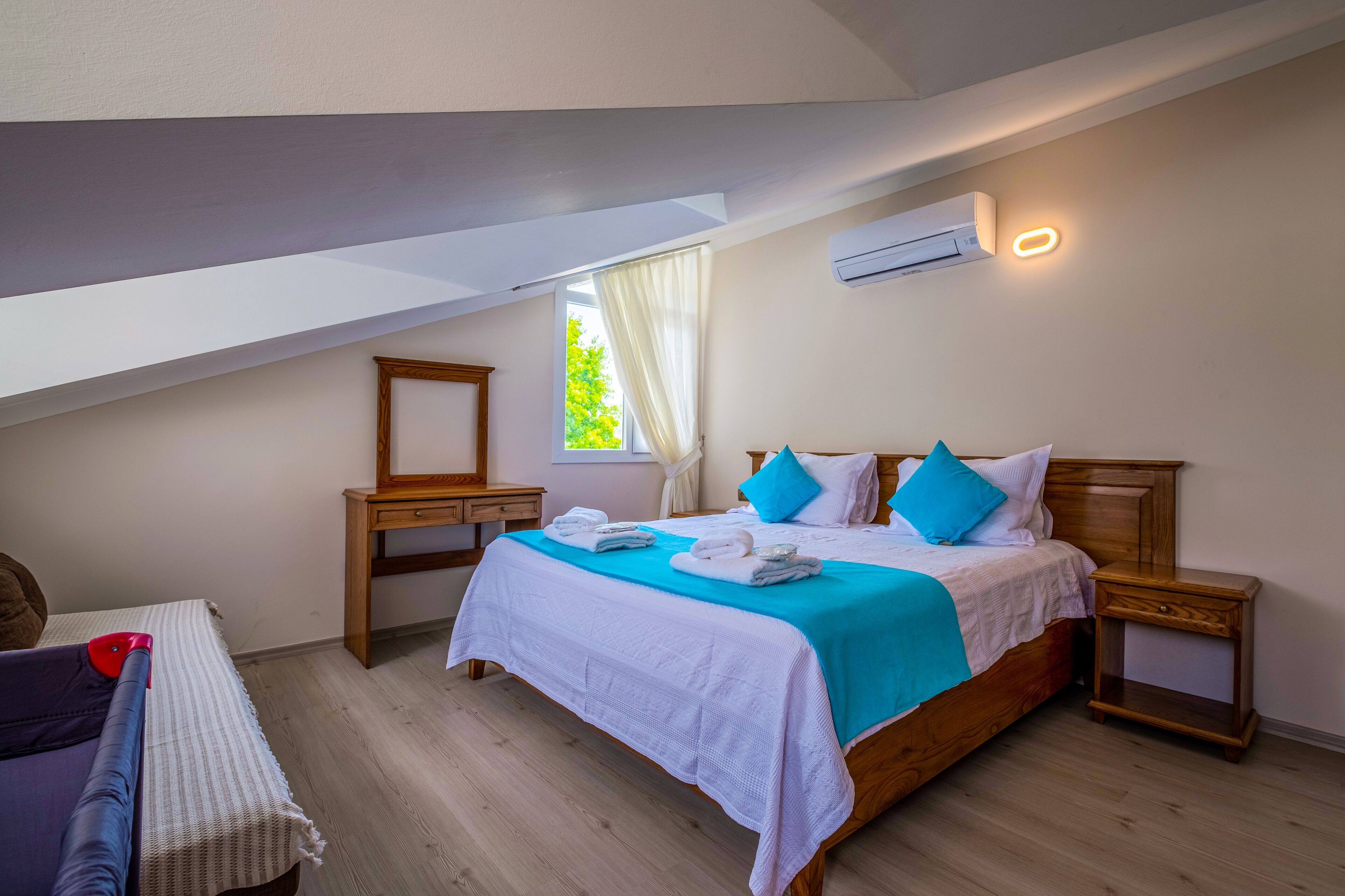 A bedroom with a sloped ceiling, a bed with a blue and white bedspread, two bedside tables, a nightstand, a window with curtains, and an air conditioning unit.