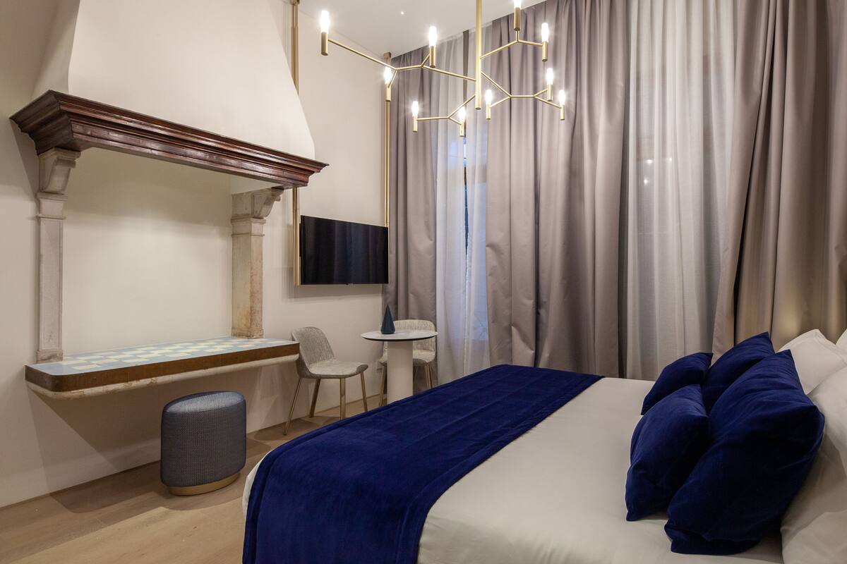 A hotel room with a bed, a small table, a chair, a TV mounted on the wall, and a window with curtains.