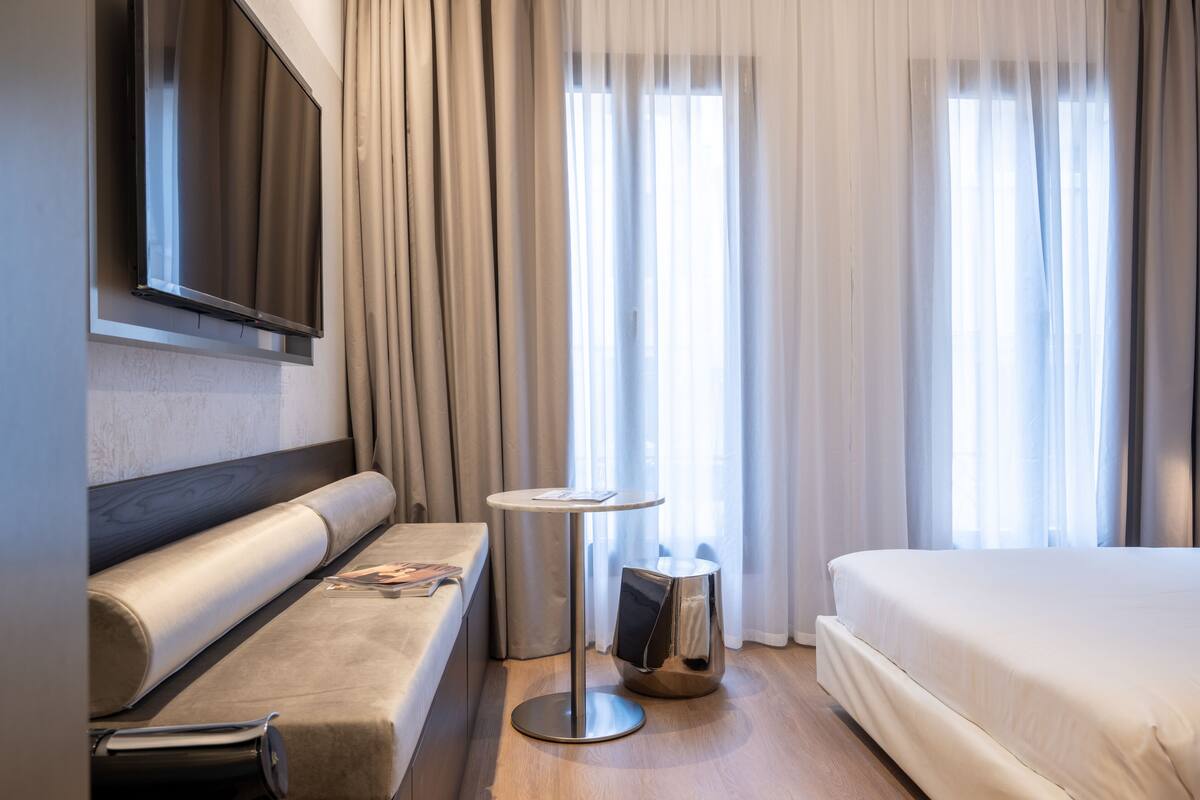 A modern hotel room with a bed, a sofa, a small round table, and a TV mounted on the wall.