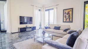 Ein modernes Wohnzimmer mit Flachbildfernseher, einer Couch, einem Couchtisch und einem großen Fenster mit Vorhängen.
