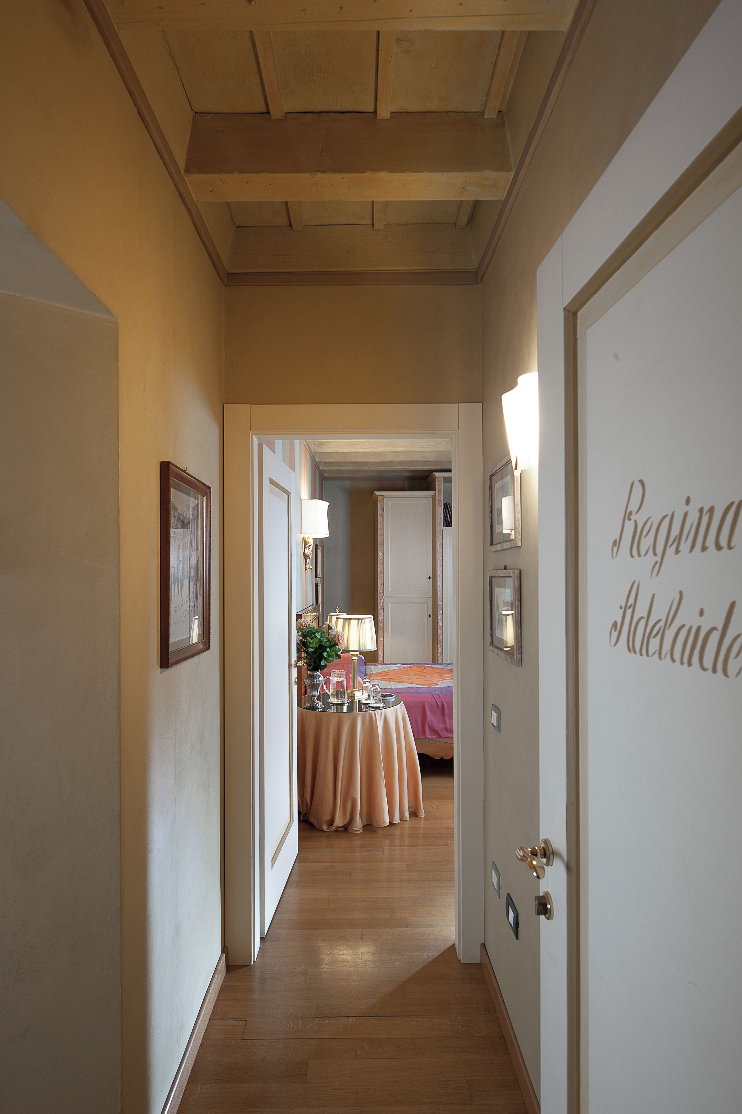 En hall som leder till ett rum med en säng, ett bord med en lampa och en dörr med texten ”Regina Adelaide”.