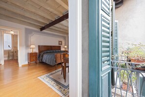 A bedroom with a wooden floor, a bed with a grey blanket, a bedside table with a lamp, and a window with blue shutters.