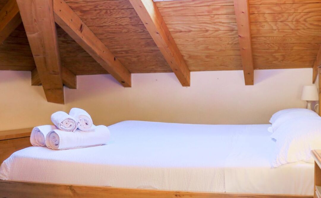A single bed with white linens, wooden beams, and a white wall.