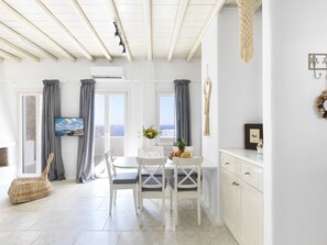 Ein heller, offener Wohnbereich mit Esstisch, Stühlen, einem Korbwäscheschrank und Blick auf das Meer durch große Fenster.
