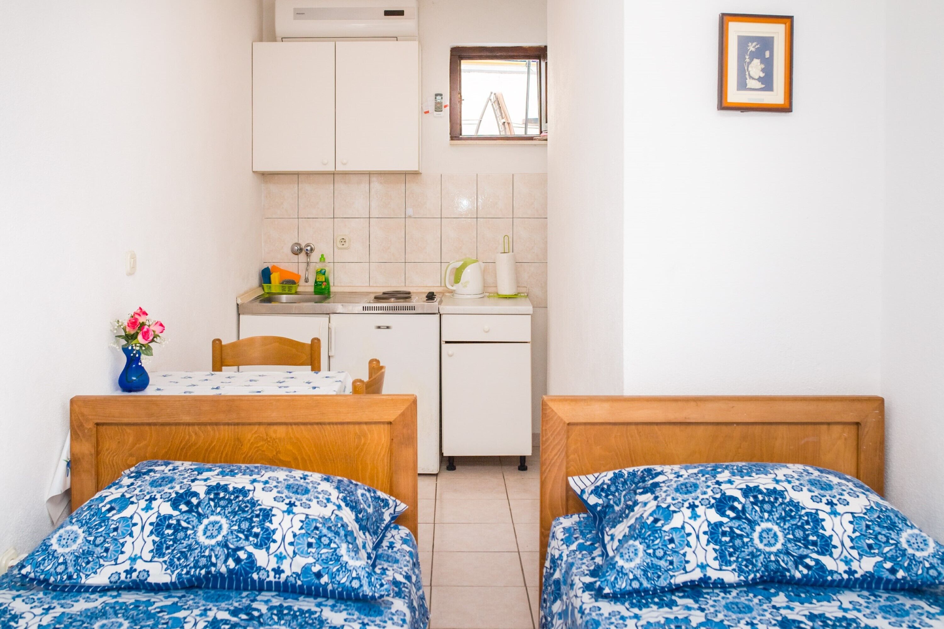 Et lille, rent værelse med to senge, en træ sengegavl, et lille bord, en hvid vask og et vindue med udsigt til en byggeplads.