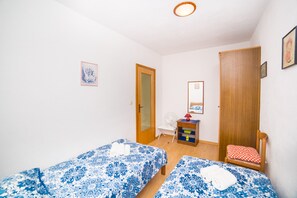 Ett litet, rent rum med två enkelsängar, en trädörr, ett litet bord med en fläkt och en stol med en röd kudde.