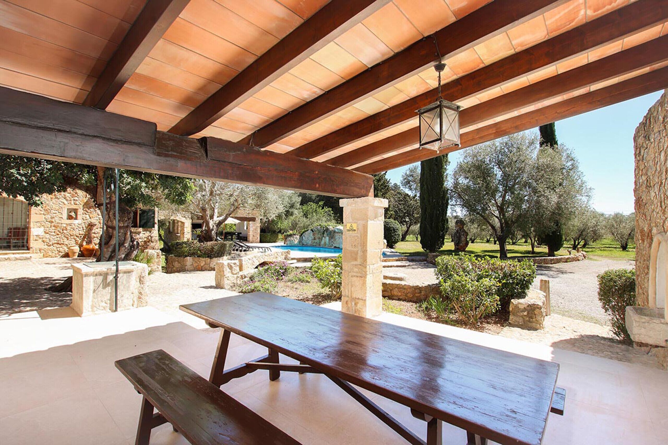 Un área de patio al aire libre con una mesa de madera y bancos, pilares de piedra y vistas a una piscina y árboles.