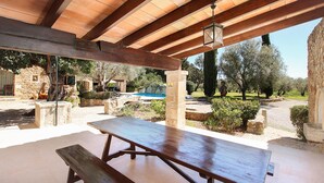 An outdoor patio area with a wooden table and benches, stone pillars, and a view of a swimming pool and trees. - Ben Sa, Finca 5StarsHome Mallorca (Puerto Pollensa)