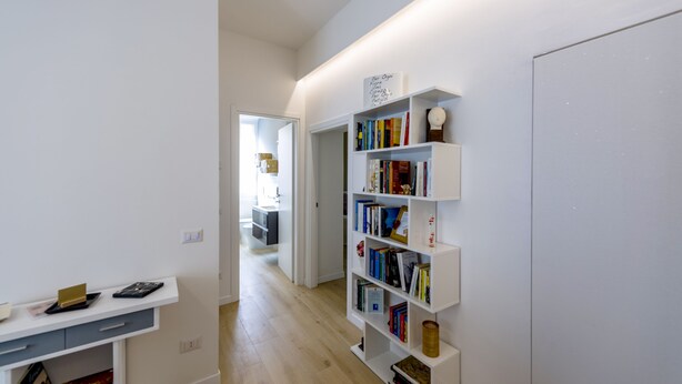 Um corredor com uma estante branca repleta de livros e objetos decorativos, uma mesa de escritório branca com um computador e livros, e uma porta que dá para outro quarto.