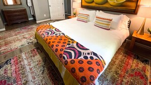 A hotel room with a large bed, colorful bedding, a fruit painting on the wall, and a wooden nightstand. - Curated for Fun with Hot Tub, Bikes, Fire Pit & More! (Kerrville)