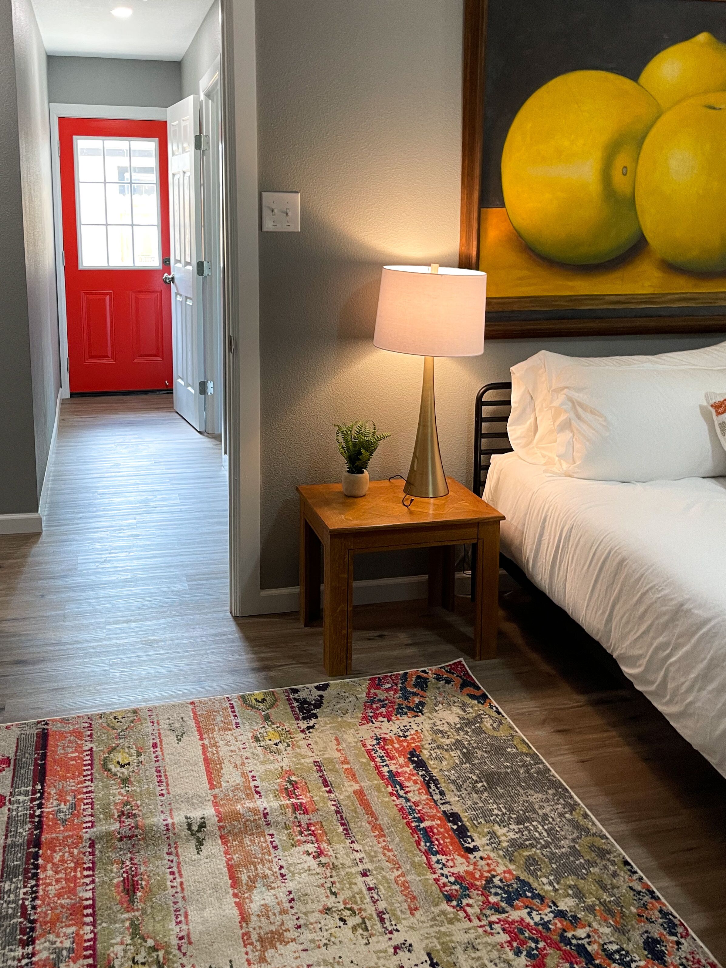 Een hotelkamer met een bed, een nachtkastje met een lamp, een kleurrijk vloerkleed, een rode deur en een schilderij van citroenen.