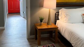 Een hotelkamer met een bed, een nachtkastje met een lamp, een kleurrijk vloerkleed, een rode deur en een schilderij van citroenen.