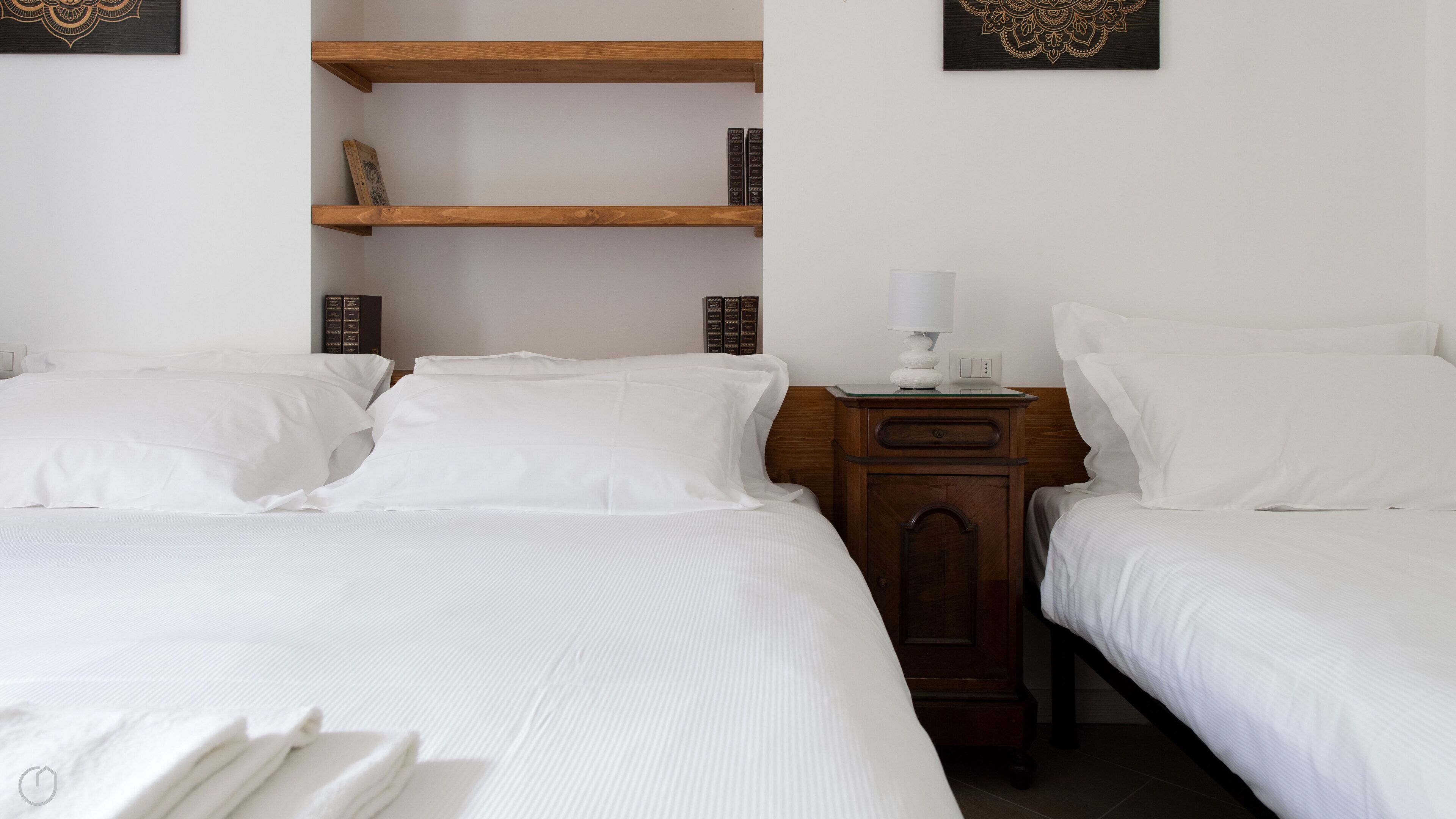 Quarto de hotel com duas camas de solteiro, uma mesa de cabeceira de madeira com abajur e uma estante com livros.